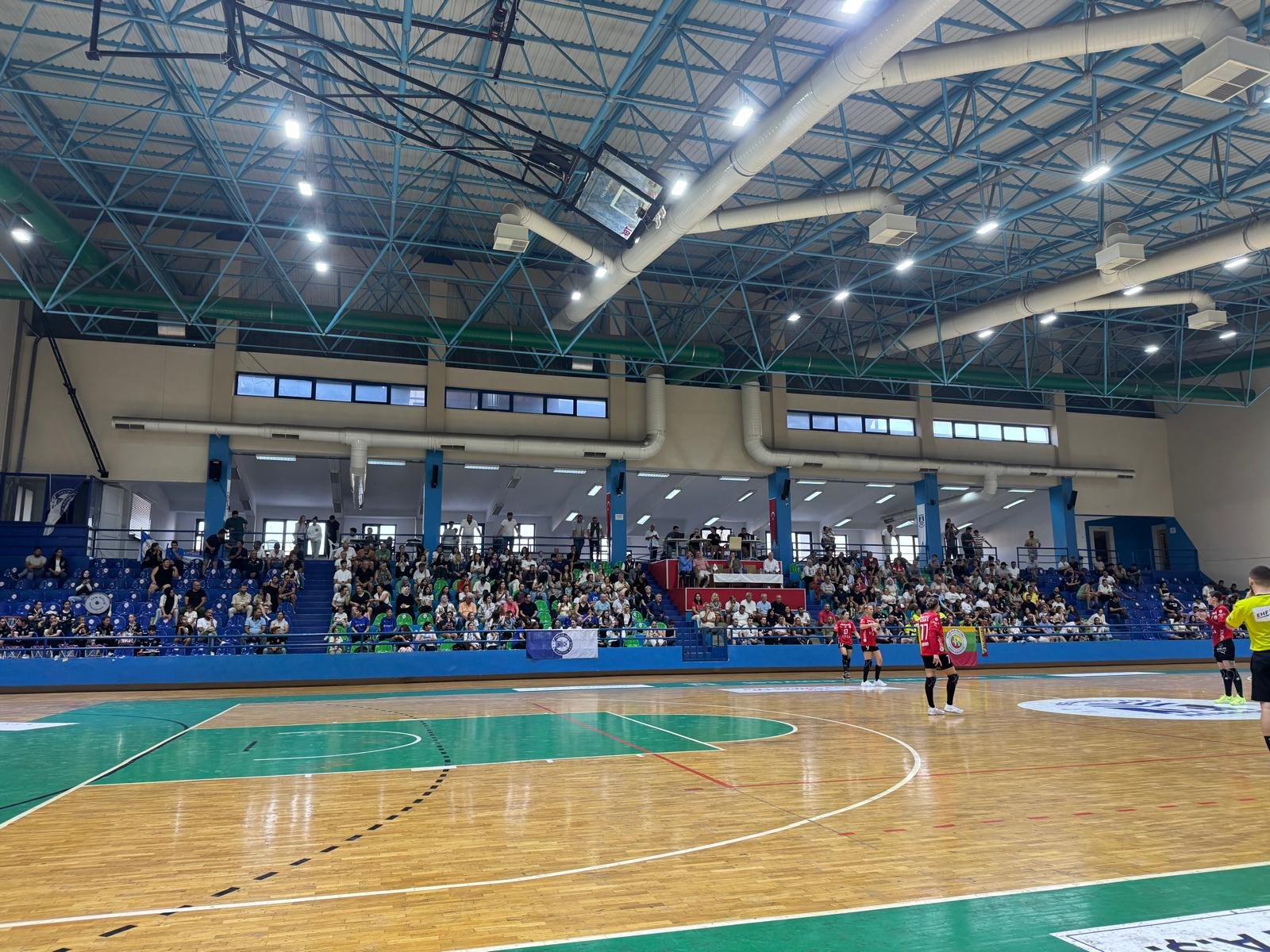 Maç öncesi spor salonu: ahşap parke, mavi-yeşil tribünler ve seyirciler